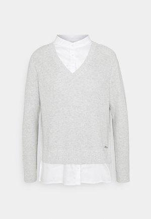 Pull gris clair à col en V en maille, superposé à une chemise blanche à boutons avec col montant, présenté sur un fond neutre.
