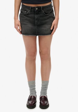 Kvinde iført sort denim miniskørt med frynset kant, grå sokker samlet sammen og bordeaux loafers, stående mod en hvid baggrund.