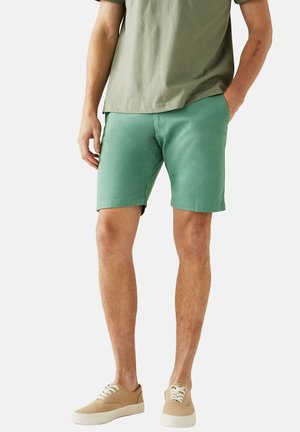 Grüne Shorts aus Baumwolle mit einem schlichten Design, geradem Schnitt und seitlichen Taschen. Kombiniert mit einem hellgrünen T-Shirt und beigen Sneakers.