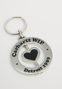 Srebrni privjesak za ključeve s crnim srcem u sredini, okrugli okvir ugraviran s "Carhartt WIP" i "Detroit 1989" u crnim slovima.