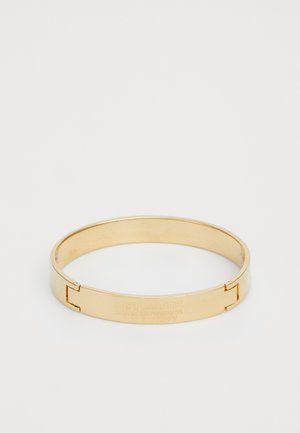 Gold metal bangle with a smooth, shiny surface. Features an engraved logo and a secure clasp closure. Circular shape, minimalist design.