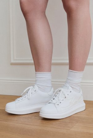 Jambes portant des chaussettes blanches mi-cheville et des baskets blanches à lacets, debout sur un sol en bois contre un lambris crème.