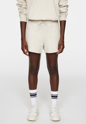 Pantaloni corti in cotone beige con cintura elastica e coulisse. Abbinati a calzini bianchi con strisce nere e sneaker di colore chiaro.