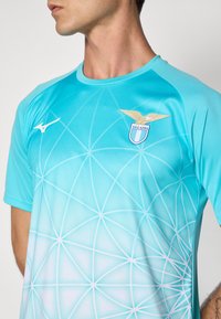 Muž oblečený v světle modré sportovní košili s bílými geometrickými liniemi, logo Mizuno na levé hrudi a emblém S.S. Lazio na pravé hrudi.