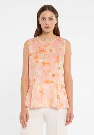 Femme portant un chemisier sans manches pêche avec un motif floral orange et blanc et un ourlet à basque, associé à un pantalon blanc, debout devant un fond blanc.