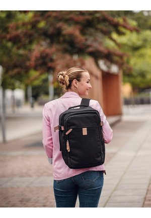 Černý batoh s hnědými akcenty, obdélníkového tvaru, s několika přihrádkami a hladkým povrchem, nošený na zádech. Růžová košile viditelná v popředí.