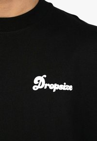 Sort bomulds T-shirt med et hvidt "Dropsize" logo i en rund, legende skrifttype. Crew neck-design med en glat tekstur.