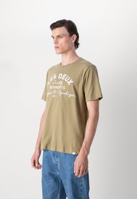 Olívovo zelené tričko z bavlny s voľným strihom, s bielym textom "LES DEUX Quality Garments" a malou etikettou s logom na leme.