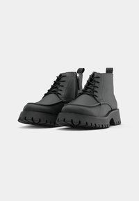 Bottines en cuir noir avec semelles épaisses à crampons, lacets sur le devant, fermetures éclair latérales et languettes à l'arrière sur un fond gris clair.