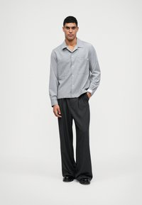Chemise grise à manches courtes et à boutons avec deux poches avant, accompagnée de pantalons larges noirs et de chaussures noires. Design simple et épuré.