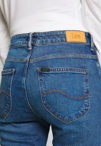 Los jeans de mezclilla azul de Lee presentan un parche de cuero marrón, bolsillos traseros con costuras en contraste y una textura desgastada que les da un aspecto usado.