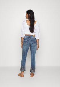 As calças de ganga azul claro com boca de sino apresentam padrões bordados na bainha, combinadas com uma blusa branca cropped e sandálias castanhas.