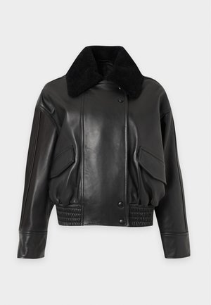 Jeune femme portant une veste en cuir noire avec un col en fourrure, un haut gris, un jean bleu à jambes larges et des chaussures marron foncé, debout devant un fond uni.