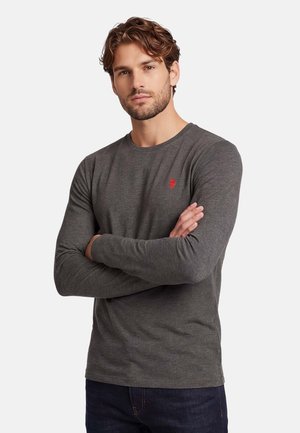 Man with light brown hair and beard wearing a fitted dark gray long-sleeve shirt with a small red emblem, arms crossed, neutral background.