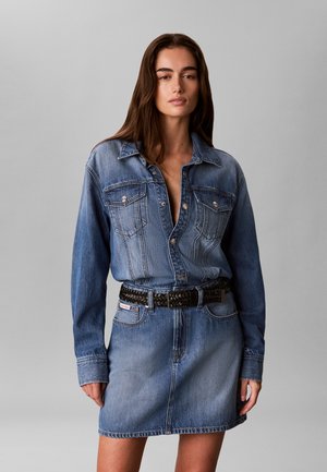 Frau mit langen braunen Haaren, die eine blaue Jeansjacke und einen passenden Jeansrock mit einem schwarzen geflochtenen Gürtel trägt, steht vor einem grauen Hintergrund.