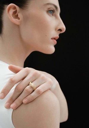 Anillo texturizado dorado sobre una mano apoyada en un hombro, mostrando un intrincado detalle y una piel suave contra un fondo negro.