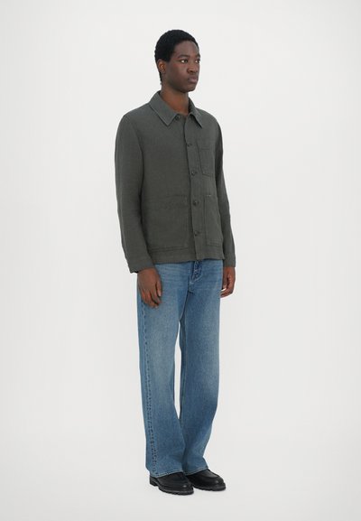 Veste boutonnée vert foncé avec un col et des poches avant, associée à un jean large bleu clair et des chaussures noires. Fond uni.