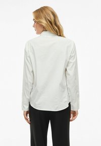 Camicia bianca a maniche lunghe con orlo curva, colletto button-down e tessuto morbido. Indossata con pantaloni neri, caratterizzati da una vestibilità comoda.