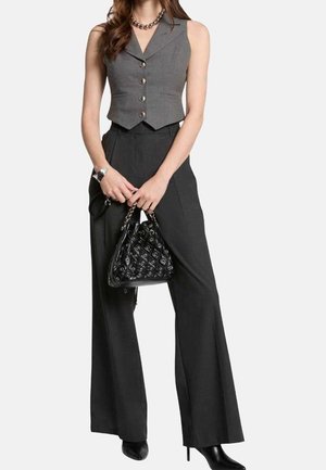 Femme portant un gilet gris sans manches boutonné, un pantalon noir à jambes larges, des bottes noires à talons, tenant un sac à main noir matelassé avec des poignées en chaîne.