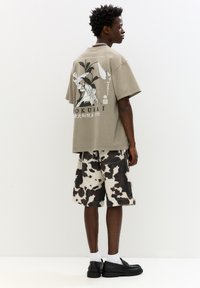 Šedé oversized tričko s grafickým designem na zadní straně, doplněné béžovými šortkami s černobílým vzorem kravské srsti. Černé boty a bílé ponožky.