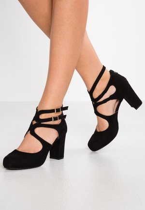 Mustat mokkakengät korkokorolla, joissa on leikkauskuvioita, nilkkaremmit kultaisilla soljilla ja paksu kanta. Neliönmuotoinen kärki vakauden takaamiseksi.