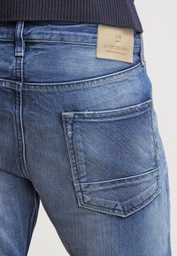 Vaqueros de mezclilla azul con un lavado desvaído, que cuentan con un bolsillo trasero, costuras amarillas y una etiqueta de cuero marrón claro con el nombre de la marca en la cinturilla.