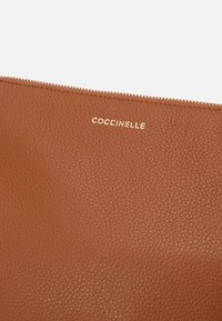 Brun läderpung med en texturerad yta, prydd med en guldfärgad varumärkeslogotyp "COCCINELLE" och ett blixtlås längs den övre kanten.