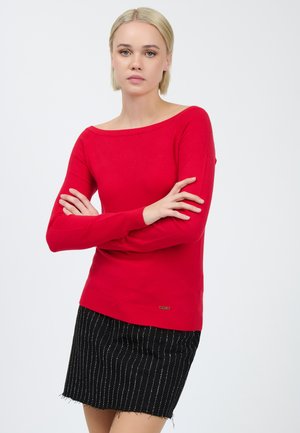 Maglione rosso a maglia con scollatura off-shoulder, maniche lunghe e polsini a costine. Abbinato a una gonna nera con righe sottili, che mette in evidenza uno stile aderente.