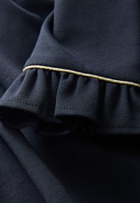 Marineblauwe stof met een gerimpelde rand, voorzien van een gouden bies. De textuur is glad en zacht, met een lichte rekbaarheid die zichtbaar is in de stof.