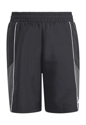 Joven con pantalones cortos deportivos negros, calcetines a rayas negros y blancos, y zapatillas negras Adidas, de pie con las manos relajadas a los costados.