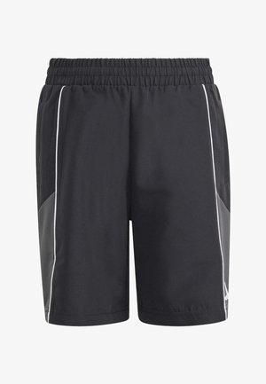 Joven con pantalones cortos deportivos negros, calcetines a rayas negros y blancos, y zapatillas negras Adidas, de pie con las manos relajadas a los costados.