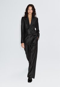 Zwart krijtstreeppak met een dubbelrijig colbert en losse broek, gemaakt van gladde stof. Afgewerkt met zwarte loafers.