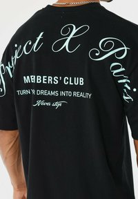 Camiseta negra usada por una persona, el texto en la parte de atrás dice "Project X Paris MEMBERS' CLUB CONVIERTE TUS SUEÑOS EN REALIDAD Nunca te detengas."