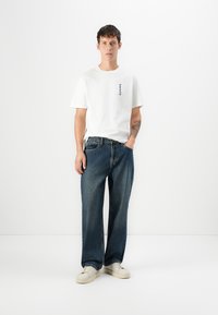 Joven de pie, llevando una camiseta blanca de manga corta, jeans holgados azules, zapatillas blancas, con una mano en el bolsillo y un tatuaje en el brazo izquierdo.