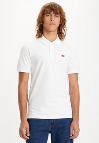 White polo shirt made of cotton, featuring a collar and short sleeves. Notable red logo on the left chest area. Classic fit design.