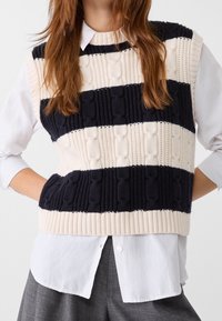 Gilet en maille avec des rayures alternées bleu marine et crème ; motifs texturés et ourlet à côtes ; porté par-dessus une chemise blanche à col.