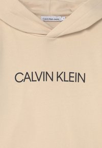 Sudadera con capucha beige hecha de tela suave, con una gran impresión negra de "CALVIN KLEIN" en el pecho y una capucha con cordón.