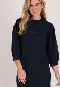 Marineblauwe sweatshirtjurk met een hoge halslijn, pofmouwen en geribbelde manchetten. Voorzien van geborduurde tekst "magique" in lichtblauw.