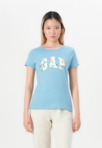 Heleheles sinine puuvillane T-särk lühikeste varrukatega, millel on valge "GAP" logo, mida ümbritsevad lillemotiivid. Pehme tekstuur, keha lähedane lõige.