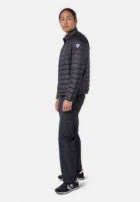Svart puffjacka med hög krage, med quiltade sektioner och en logopatch på vänster axel. Bärs med mörka byxor och svarta sneakers.