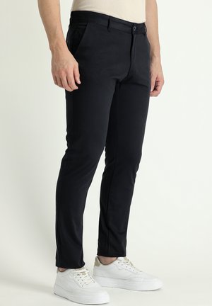 Zwarte slim-fit broeken met een gladde textuur, voorzien van zakken en een knoopsluiting. Gecombineerd met witte sneakers.