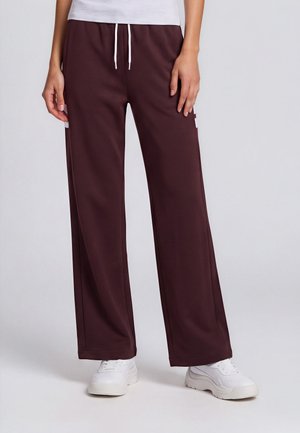 Personne portant un pantalon ample bordeaux avec un cordon blanc et des baskets blanches, debout devant un fond uni clair.