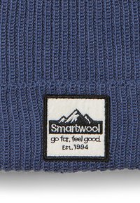 Bonnet en tricot bleu foncé avec une texture côtelée. Présente une étiquette avec des montagnes blanches et le logo "Smartwool". Le texte sur l'étiquette inclut "allez loin, ressentez-vous bien, Etab. 1994."