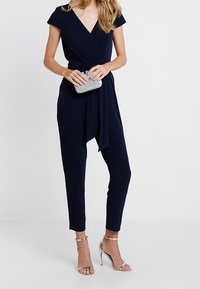 Mono azul marino con escote en V profundo, mangas cortas y cintura anudada. Combinado con sandalias de tacón alto plateadas y un clutch brillante.