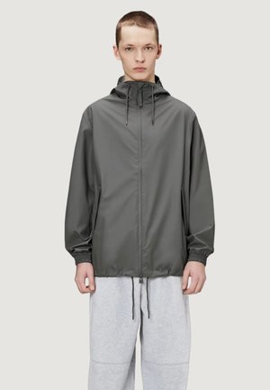 Joven con chaqueta con capucha y cremallera de color gris oscuro con cordones y pantalones deportivos gris claro, de pie frente a un fondo liso.
