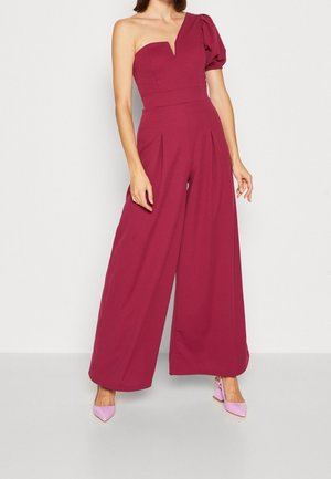 Femme portant une combinaison magenta sans manches avec une manche bouffante et un pantalon pattes d'éléphant, chaussée de talons roses à bouts pointus, debout contre un mur blanc.