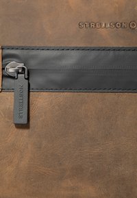 Borsa in pelle marrone con superficie texturizzata, dotata di una striscia in nylon nera, cerniera in metallo e un'etichetta stampata con "STRELLSON."