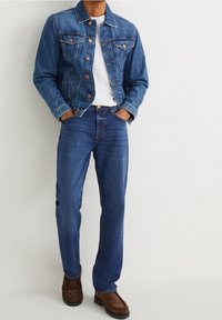 Man wearing blue denim jacket and jeans, white t-shirt, and brown suede shoes, standing with hands in pockets against white background.