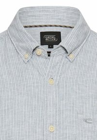 Jasnoszara koszula zapinana na guziki z cienkimi białymi paskami, beżowymi guzikami, kołnierzykiem oraz małym logo wyszywanym na kieszeni na piersi.