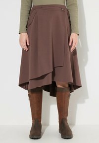 Bruine wikkelrok met een asymmetrische zoom, gemaakt van zacht materiaal; heeft zijzakken en een decoratieve knop. Gecombineerd met bruin-kneehoog laarzen.
